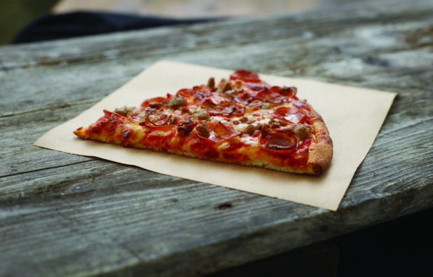 A Jumbo Slice of Meat Lovers Pizza with Pepperoni, Sausage, Bacon and Ham on Parchment Paper on an Aged Wood Background - Variation