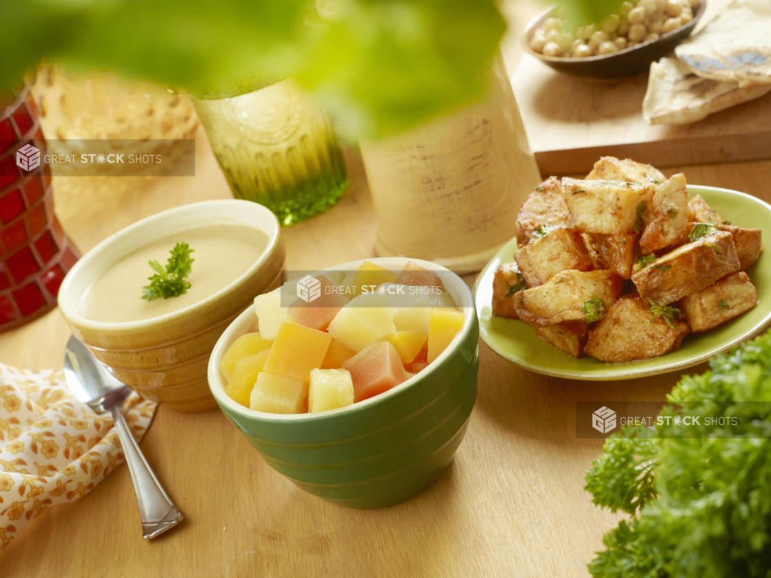 Assortment of Side Dishes - Roasted Garlic Potatoes, Tropical Fruit Salad and a Bowl of Lentil Soup - on a Wooden Table with Fresh Parsley and Other Ingredients in an Indoor Setting