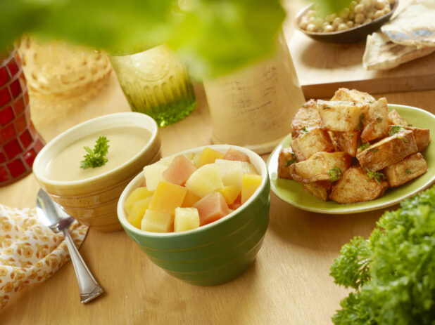 Assortment of Side Dishes - Roasted Garlic Potatoes, Tropical Fruit Salad and a Bowl of Lentil Soup - on a Wooden Table with Fresh Parsley and Other Ingredients in an Indoor Setting