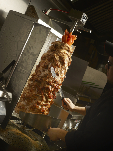 Chicken Shawarma on a Rotating Vertical Spit with a Chef Shaving Slices of Meat into a Stainless Steel Scoop in a Restaurant Food Preparation Setting