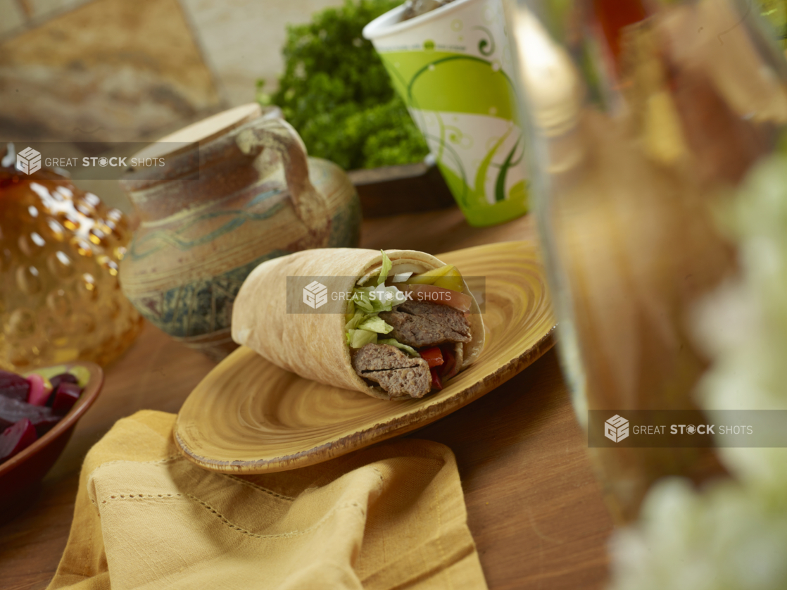 Beef Kebab Pita Wrap with Pickled Veggies and Fresh Vegetables on a Round Ceramic Dish with a Cold Drink on a Wooden Table in an Indoor Setting