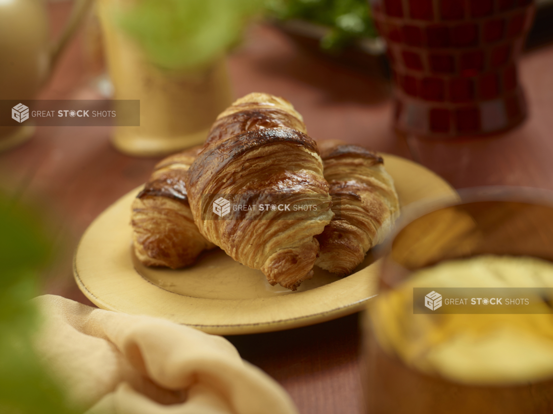 A Trio of Fresh Baked Butter Croissants on a Round Yellow Plate on a Wooden Table in an Indoor Setting