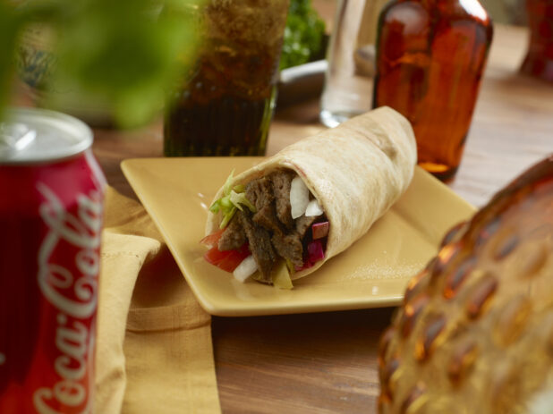 Beef Shawarma Pita Wrap with Pickled Veggies and Fresh Vegetables and a Can of Soda on a Rectangular Ceramic Dish on a Wooden Table in an Indoor Setting