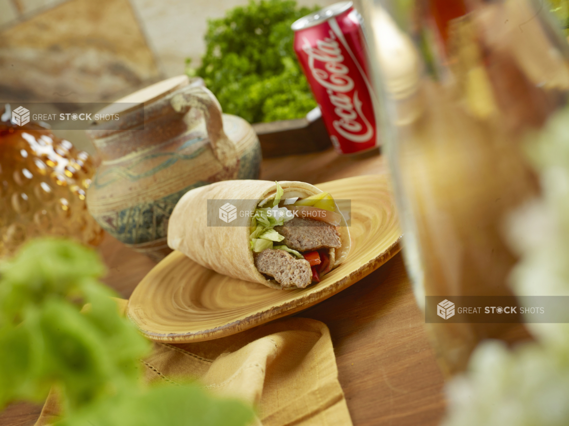 Beef Kebab Pita Wrap with Pickled Veggies and Fresh Vegetables on a Round Ceramic Dish with a Can of Coke on a Wooden Table in an Indoor Setting