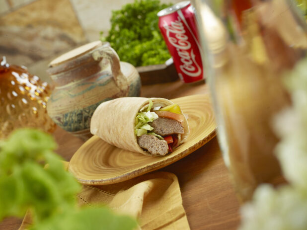 Beef Kebab Pita Wrap with Pickled Veggies and Fresh Vegetables on a Round Ceramic Dish with a Can of Coke on a Wooden Table in an Indoor Setting