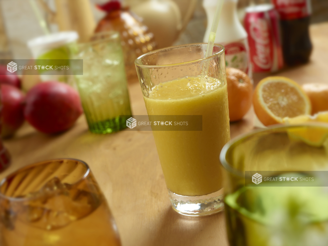 Glass of Fresh Squeezed Orange Juice, Ice Water, Fountain Soda, Can and Bottle of Coke and Yogurt Drinks on a Wooden Table with Colourful Glassware in an Indoor Setting