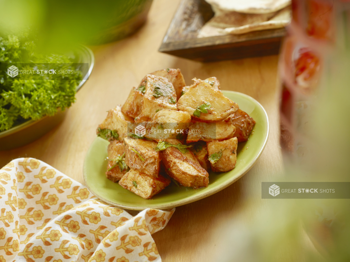 A Round Green Plate of Roasted Garlic Potatoes with Cilantro and Chilli Spices on a Wooden Table in an Indoor Setting