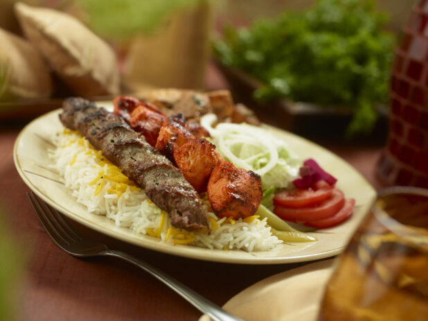 A Middle Eastern Style Dinner Combo Plate with Beef and Chicken Kebabs, Garlic Potatoes and Fresh Veggies on a Round Dinner Plate on a Wooden Table in an Indoor Setting
