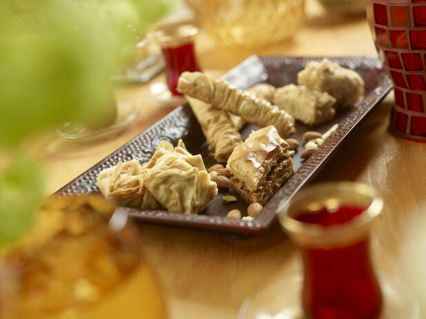 A Brown Rectangular Ceramic Platter with Baklava Pastry in Varying Sizes and Shapes on a Wooden Table in an Indoor Setting