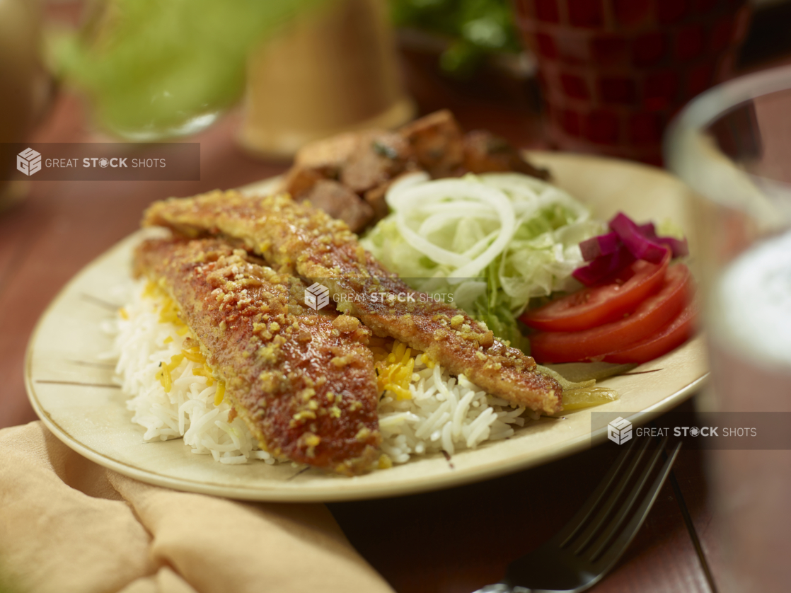 A Middle Eastern Style Dinner Combo Plate with Seasoned Baked Fish, Garlic Potatoes and Fresh Veggies on a Round Dinner Plate on a Wooden Table in an Indoor Setting