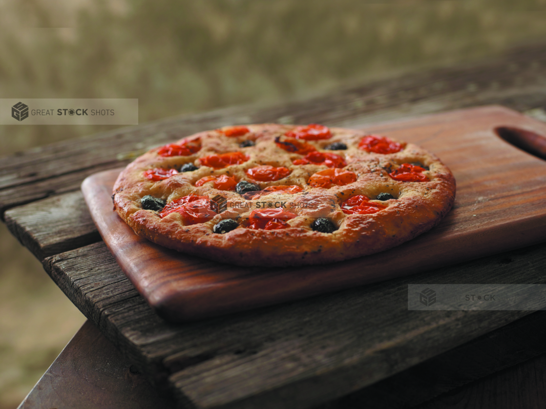 A Roasted Tomato and Black Olive Round Focaccia Bread on a Wooden Cutting Board on a Wooden Table Against a Brown Canvas Background