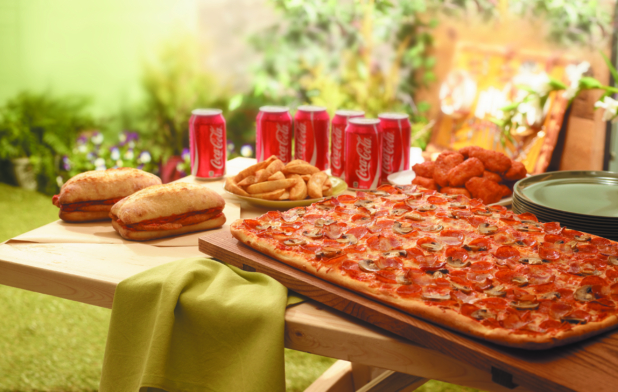 A Pizza Party with a Large Sicilian Pizza, Boneless Wings, Potato Wedges, Italian Sandwiches and a 6-Pack of Coca-Cola Cans on a Picnic Table in an Outdoor Garden Setting