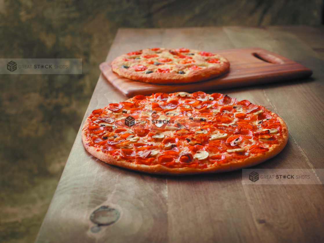 Large Whole Canadian Pizza with Pepperoni, Mushrooms and Bacon Toppings on a Wooden Table with a Tomato and Olive Focaccia Bread against a Brown Canvas Background