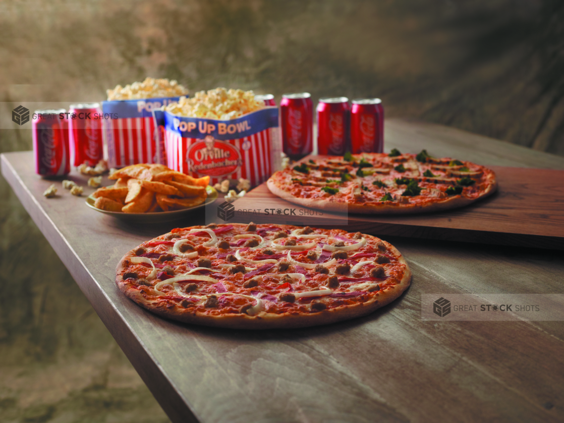 Movie Night Special with Two 3-Topping Pizzas, Microwave Popcorn, Potato Wedges and Cans of Coca-Cola on a Wooden Table Against a Brown Canvas Background