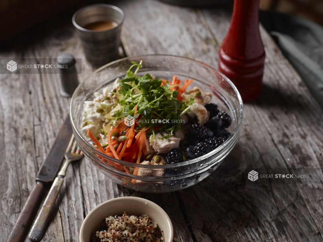 Glass Bowl with Pea Shoots, Julienne Carrots, Grilled Chicken, Tofu and Berries on an Aged Wooden Surface with a Cup of Dressing and Quinoa