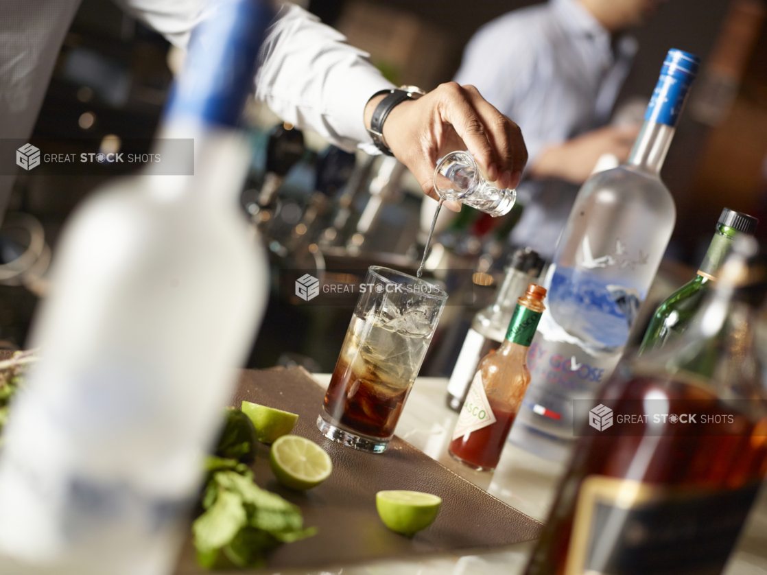 Bartender's Hands Pouring a Shot of Vodka into a Glass of Cola with Ice Cubes in a Restaurant Bar Setting with Bottles of Assorted Alcohol