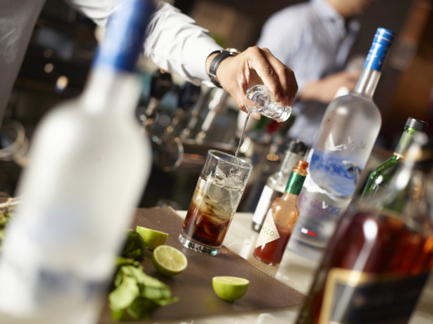 Bartender's Hands Pouring a Shot of Vodka into a Glass of Cola with Ice Cubes in a Restaurant Bar Setting with Bottles of Assorted Alcohol