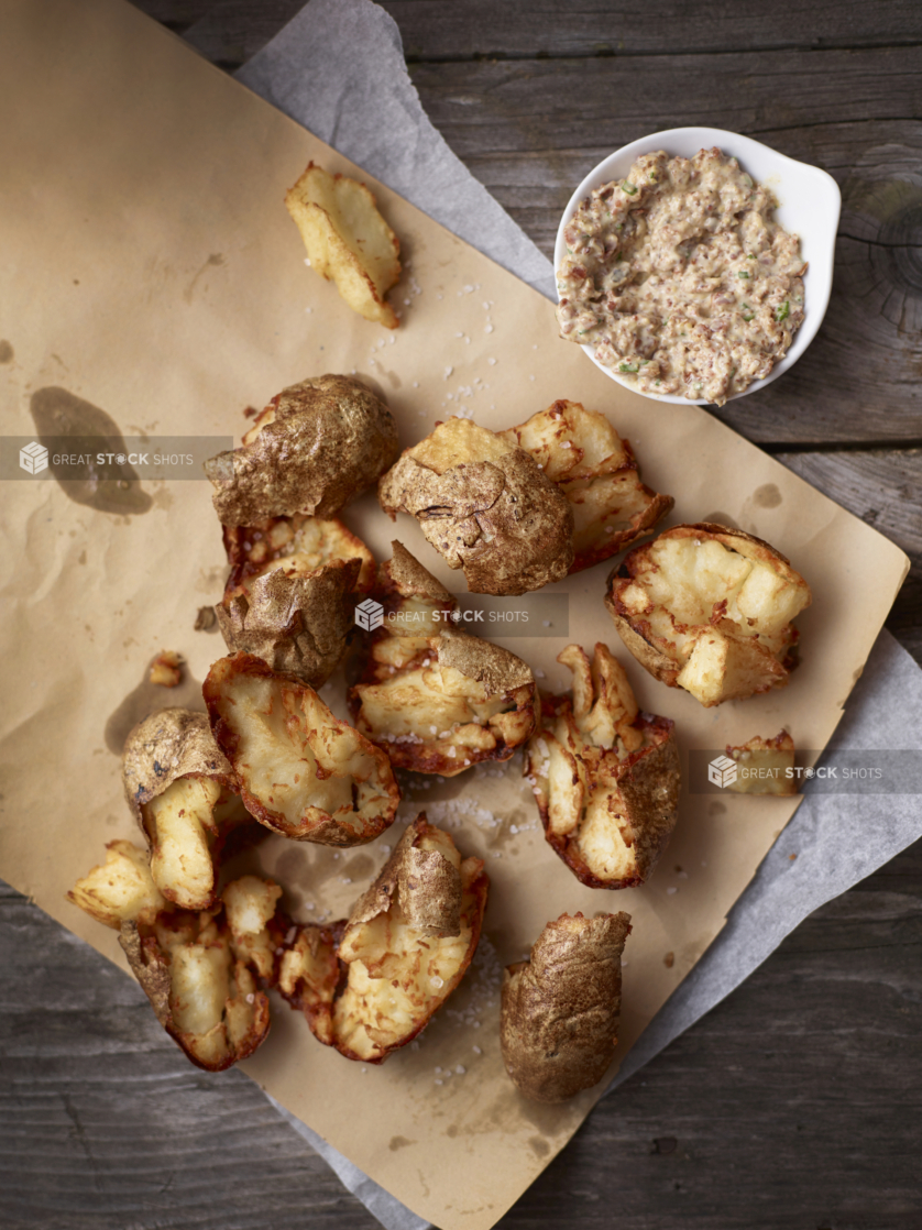 Overhead View of Smashed Baked Potatoes on Kraft and Parchment Paper on an Aged Wooden Surface with a Side Bowl of Sour Cream, Bacon and Onions Dip