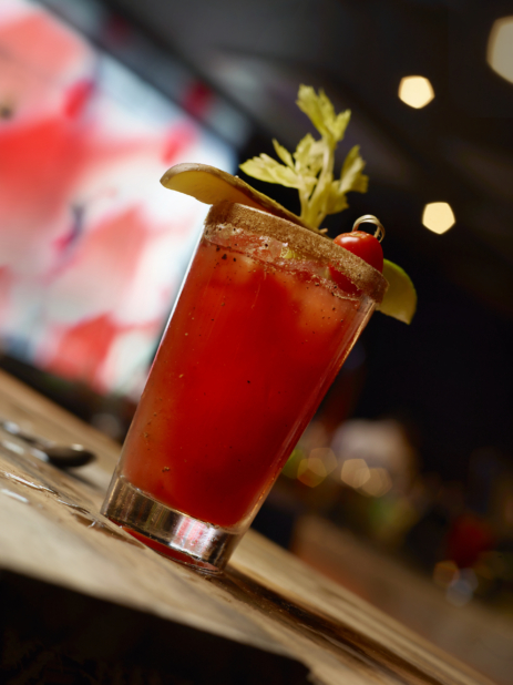 Close Up of a Glass of Bloody Caesar with Celery Salt Rim, Lime Wedge, Cherry Tomato, Celery Stalk and Pickle Spear Garnish on a Wooden Table, Viewed from Below