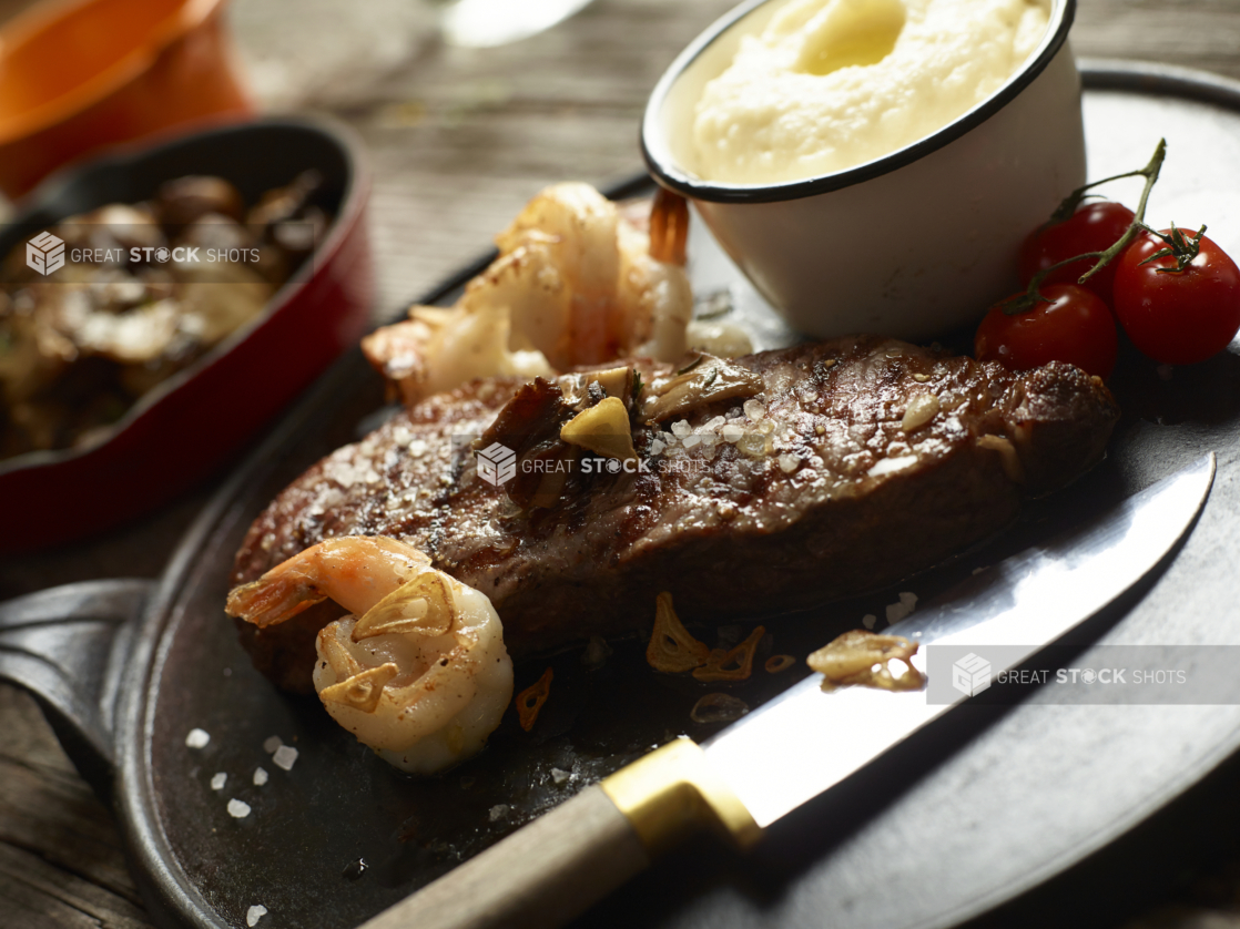 Cast Iron Platter with Surf and Turf Dinner of Sautéed Shrimp and Grilled Sirloin Steak with a Side Bowl of Buttery Mashed Potatoes and Cherry Tomatoes on an Aged Wood Surface