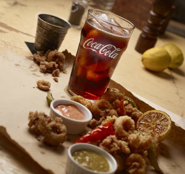 Calamari Piled on Kraft Paper with Dipping Sauces in Ramekins, a Roasted Lemon Half and a Glass of Coca-Cola on White Painted Wooden Table