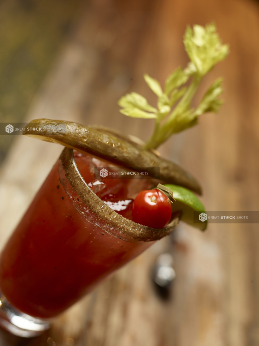 Glass of Bloody Caesar with Celery Salt Rim, Lime Wedge, Cherry Tomato, Celery Stalk and Pickle Spear Garnish on an Aged Wooden Surface