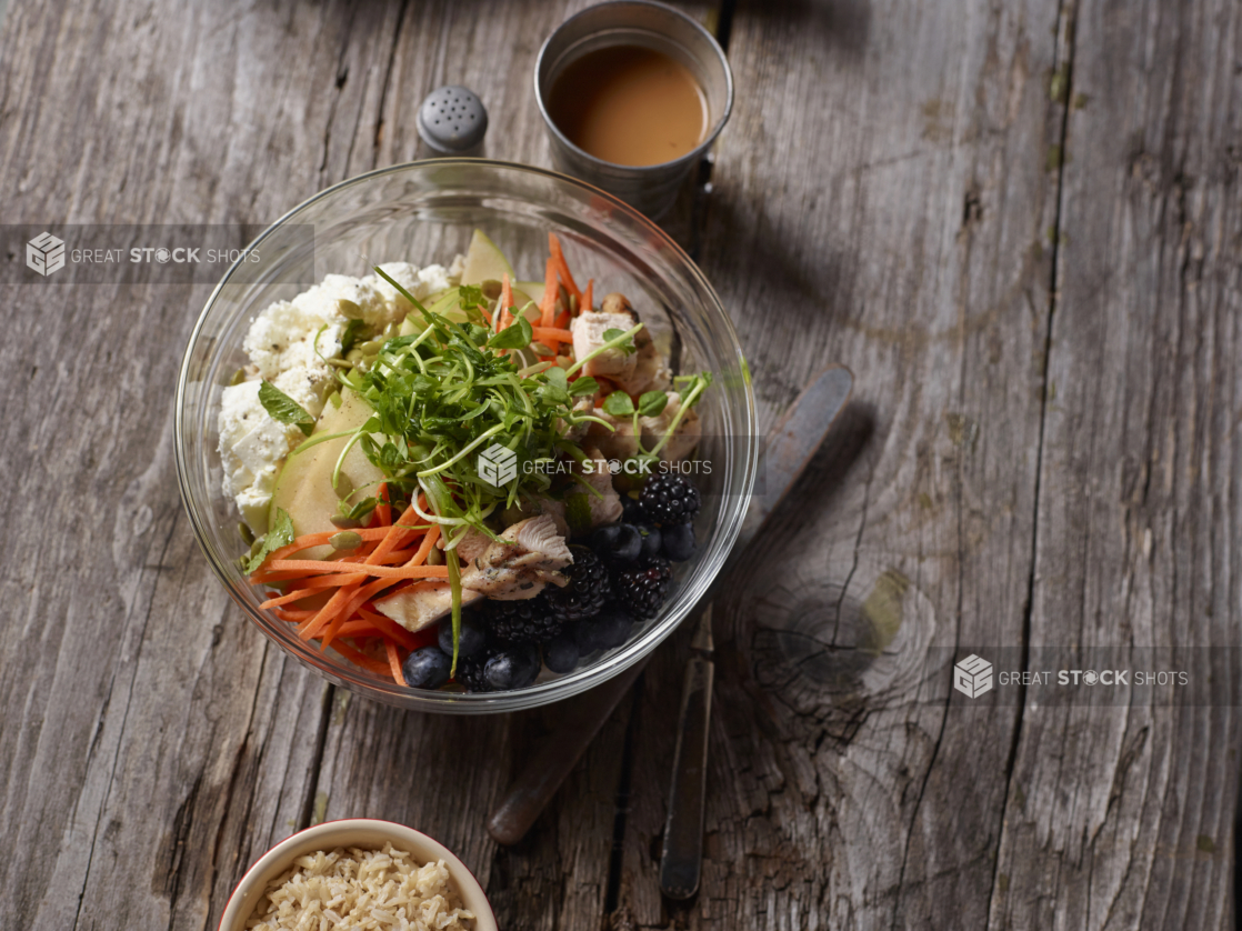 Overhead View of a Glass Bowl with Pea Shoots, Julienne Carrots, Grilled Chicken, Tofu and Berries on an Aged Wooden Surface with a Cup of Dressing and Brown Rice