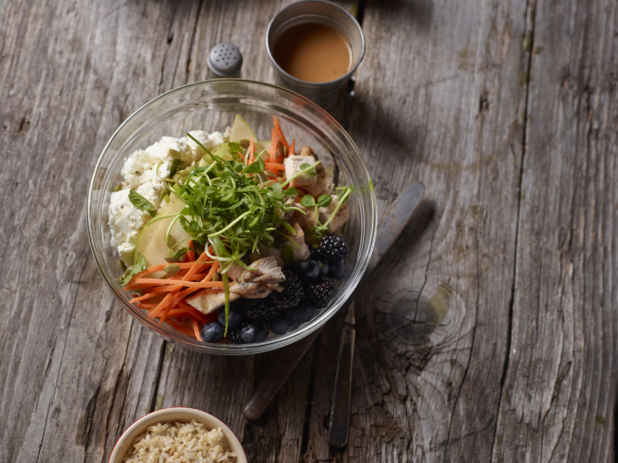 Overhead View of a Glass Bowl with Pea Shoots, Julienne Carrots, Grilled Chicken, Tofu and Berries on an Aged Wooden Surface with a Cup of Dressing and Brown Rice