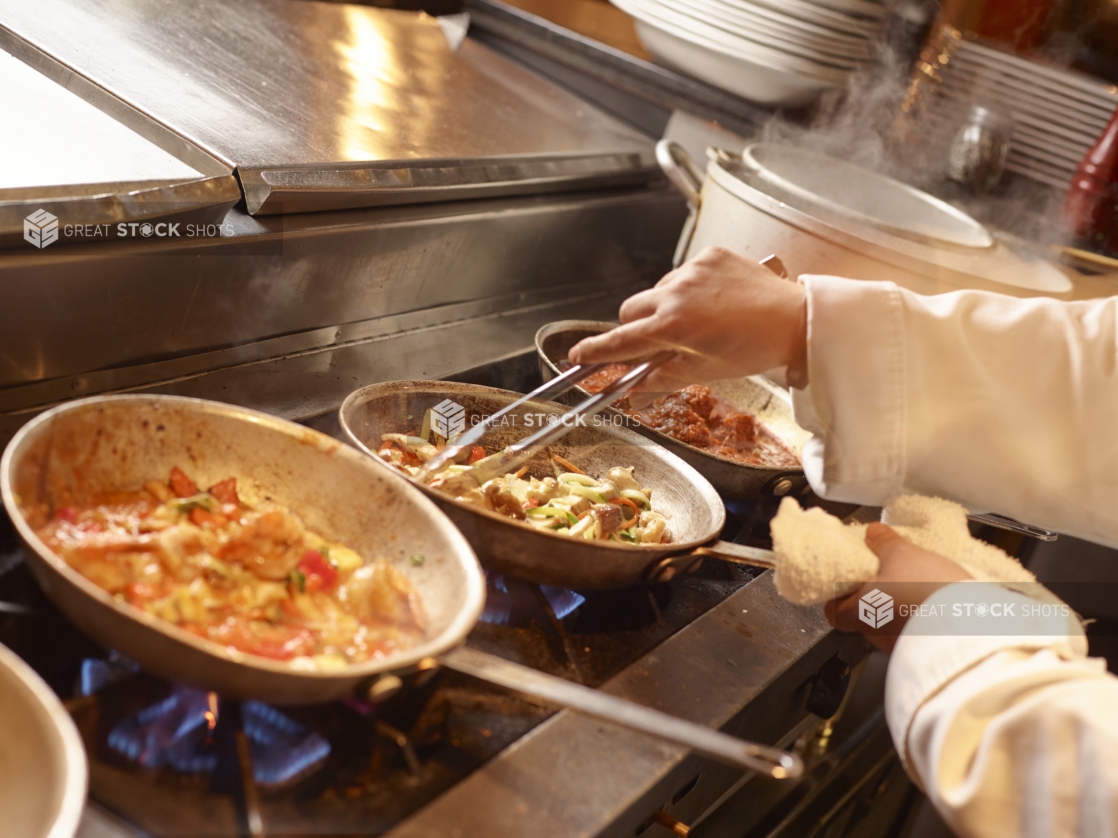 Back of House Kitchen Setting with Chef’s Hands Cooking Vegetables in a Frying Pan with Tongs Over a Gas Fire Stove