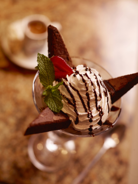 Overhead View of a Mini Parfait in Glass Cup with Whipped Cream, Brownie Wedges and Chocolate Drizzle on a Wooden Surface in an Indoor Restaurant Setting