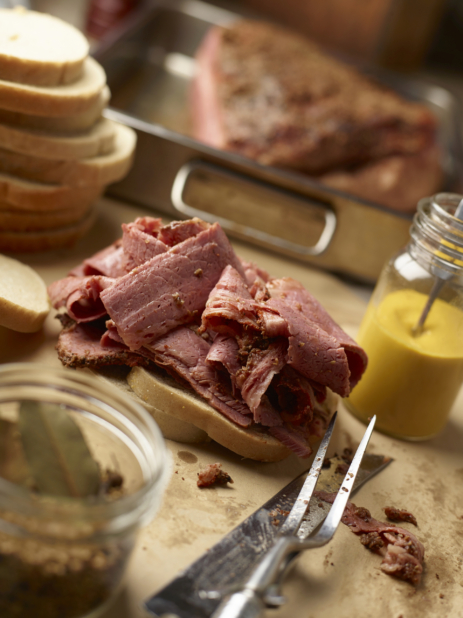 Pastrami Sandwich Being Made with Layers of Sliced Pastrami Deli Meat on White Tye Bread with a Jar of Home-made Mustard and Meat Caring Utensils