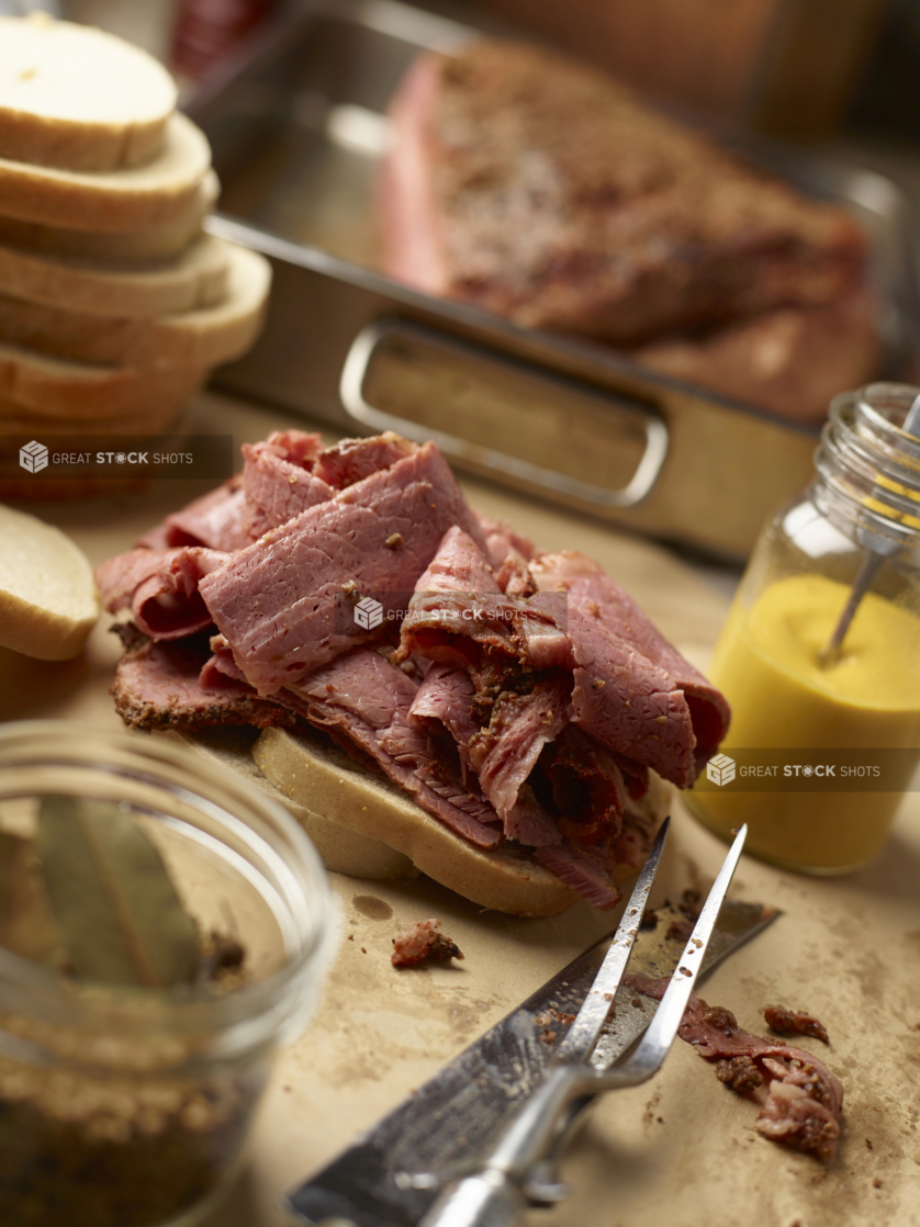 Pastrami Sandwich Being Made with Layers of Sliced Pastrami Deli Meat on White Tye Bread with a Jar of Home-made Mustard and Meat Caring Utensils