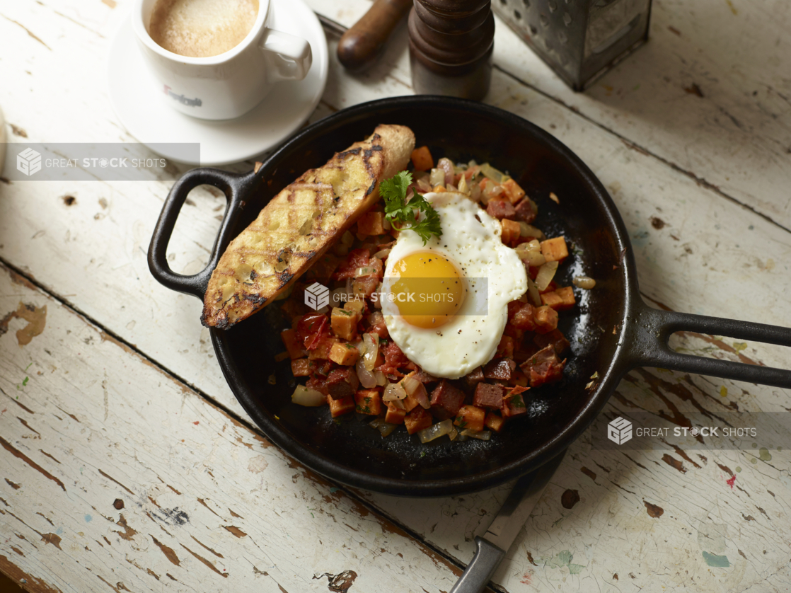 Roasted Veggie, Sweet Potatoes and Sausage Hash in a Cast Iron Skillet with a Fried Sunny Side up Egg and a Grilled Buttered Baguette on a White Pained Wood Surface