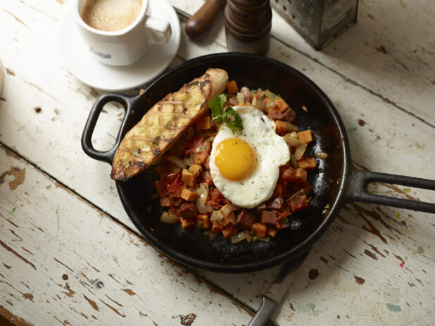 Roasted Veggie, Sweet Potatoes and Sausage Hash in a Cast Iron Skillet with a Fried Sunny Side up Egg and a Grilled Buttered Baguette on a White Pained Wood Surface