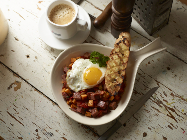 Roasted Veggie, Sweet Potatoes and Sausage Hash in a Ceramic Bowl with a Fried Sunny Side Up Egg and a Grilled Buttered Baguette on a White Pained Wood Surface