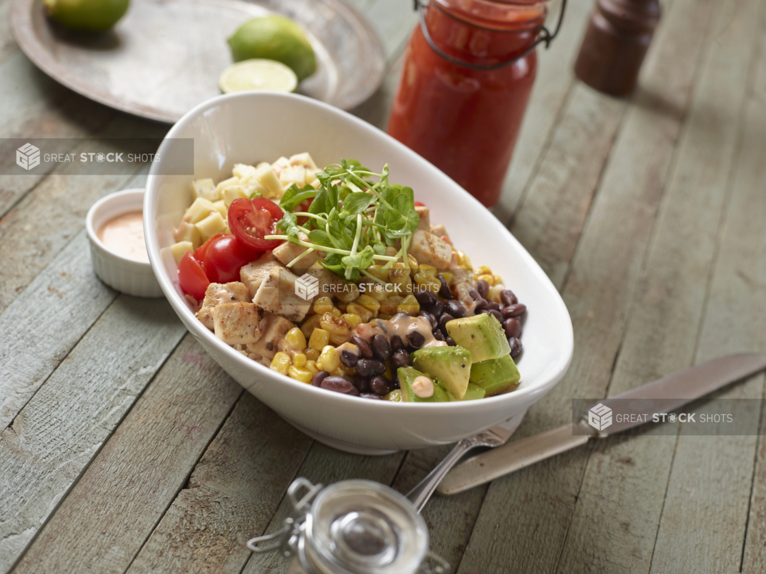 Large White Oval Bowl with Cubed Grilled Chicken, Corn, Black Beans, Avocado, Cherry Tomatoes, Cheese and Pea Shoots on a Weathered Wooden Surface in an Indoor Setting