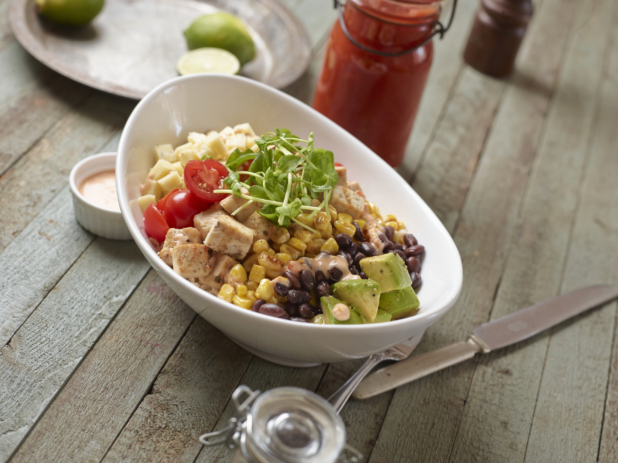 Large White Oval Bowl with Cubed Grilled Chicken, Corn, Black Beans, Avocado, Cherry Tomatoes, Cheese and Pea Shoots on a Weathered Wooden Surface in an Indoor Setting