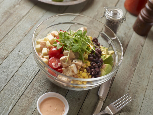 Large Glass Bowl with Cubed Grilled Chicken, Corn, Black Beans, Avocado, Cherry Tomatoes, Cheese and Pea Shoots on a Weathered Wooden Surface in an Indoor Setting