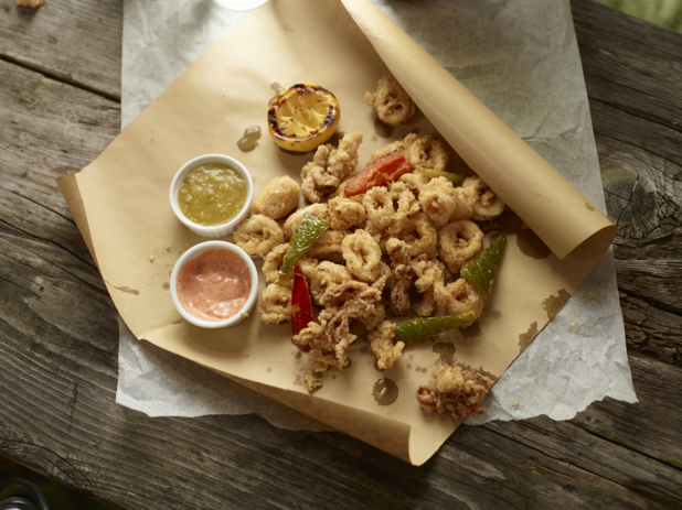 Overhead View of Calamari Piled on Kraft Paper with Dipping Sauces in Ramekins, a Roasted Lemon Half on Aged Wooden Table