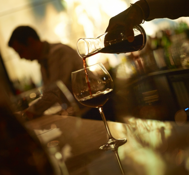 Bartender's Hands Pouring a Glass Carafe of Red Wine into a Wine Glass on a Wooden Bar Counter in a Restaurant Setting