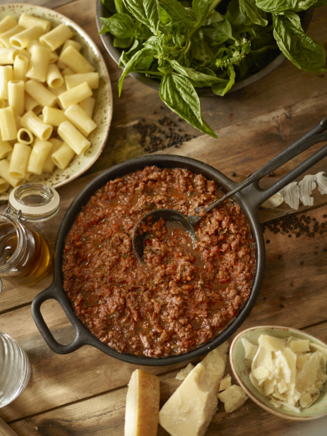 Overhead View of a Cast Iron Pot with Hearty Homemade Bolognese Sauce with Rigatoni Pasta, Fresh Basil Leaves and Shaved Parmigiana-Reggiano Cheese on a Wooden Surface