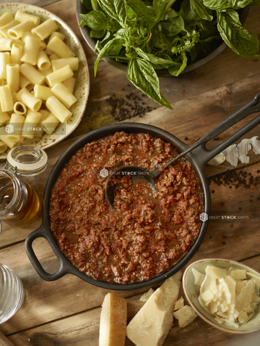 Overhead View of a Cast Iron Pot with Hearty Homemade Bolognese Sauce with Rigatoni Pasta, Fresh Basil Leaves and Shaved Parmigiana-Reggiano Cheese on a Wooden Surface