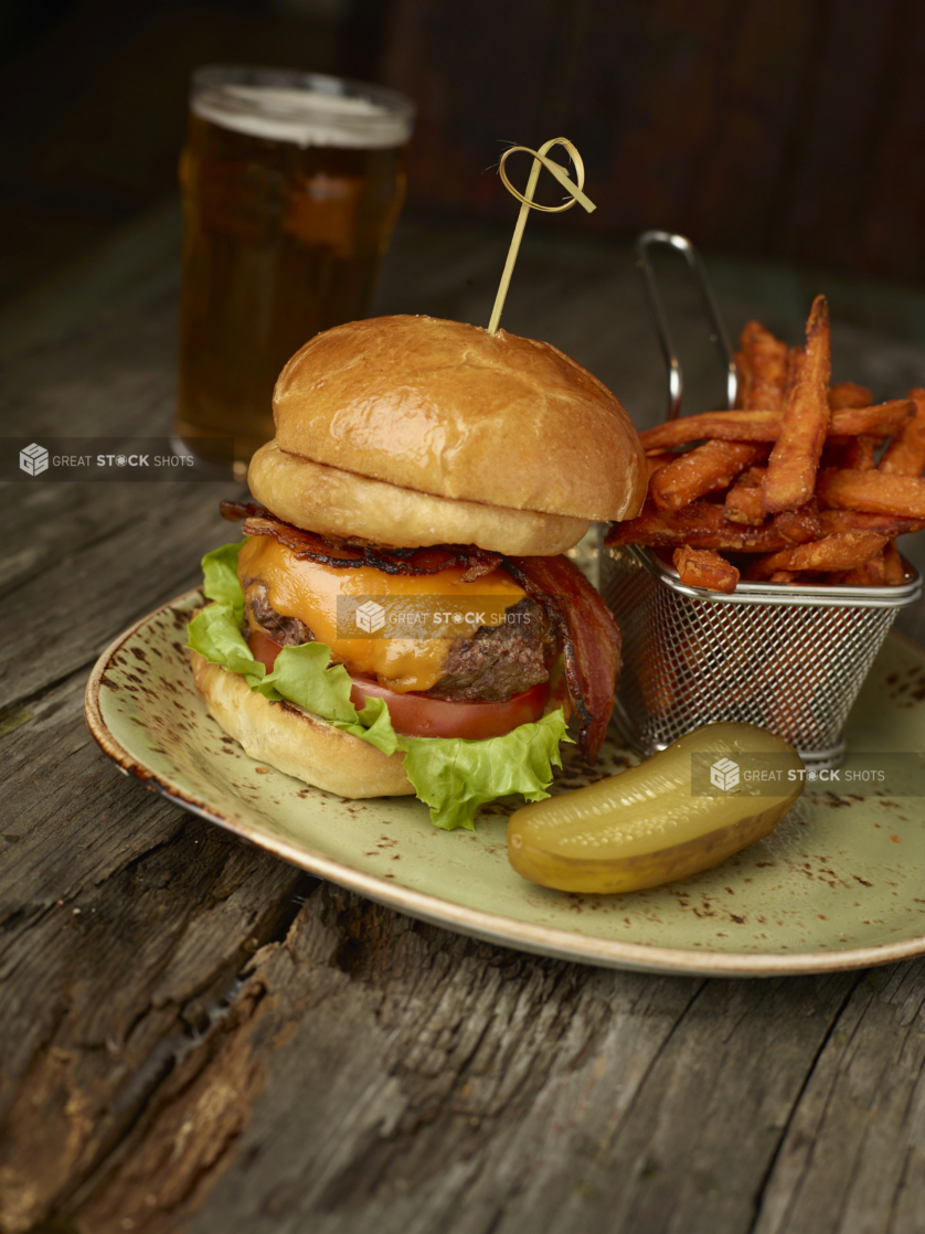 Gourmet Cheeseburger with Bacon, Lettuce and Tomato with Sweet Potato Fries and Half a Pickle on a Ceramic Dish with a Pint of Draught Beer on an Aged Wooden Table