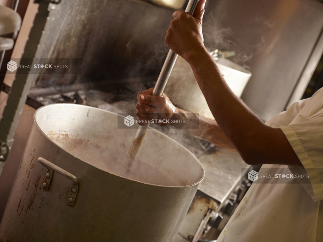 Chef's Hands Mixing Food in a Large Commercial Cooking Pot with Steam Swirling Around in a Back Kitchen Setting of a Restarant