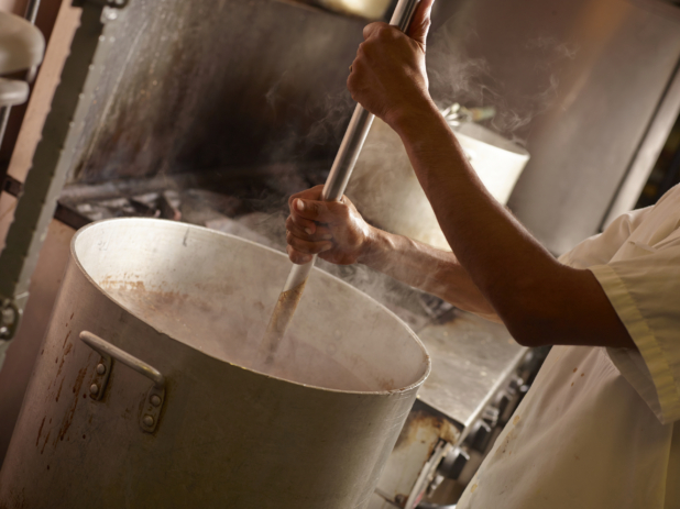 Chef's Hands Mixing Food in a Large Commercial Cooking Pot with Steam Swirling Around in a Back Kitchen Setting of a Restarant