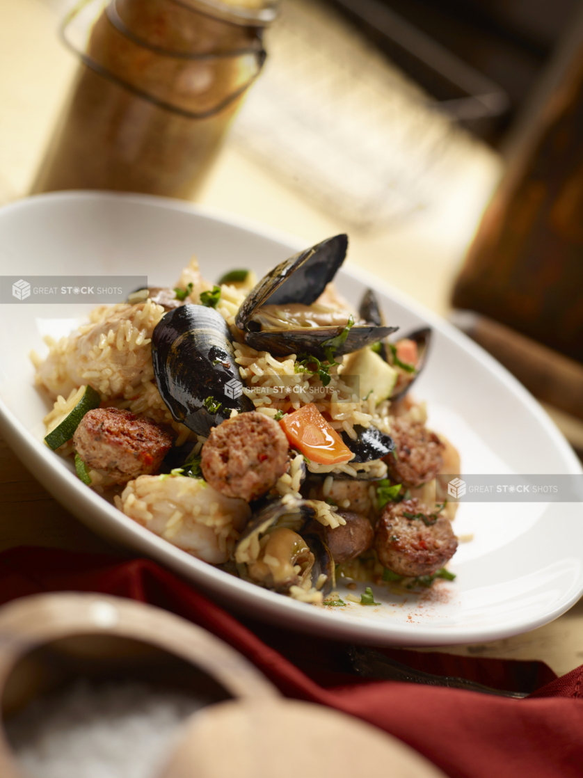 Round White Dish of Seafood Paella on a White Painted Wooden Table with a Red Napkin and Wooden and Glass Kitchen Items with Bokeh Effect