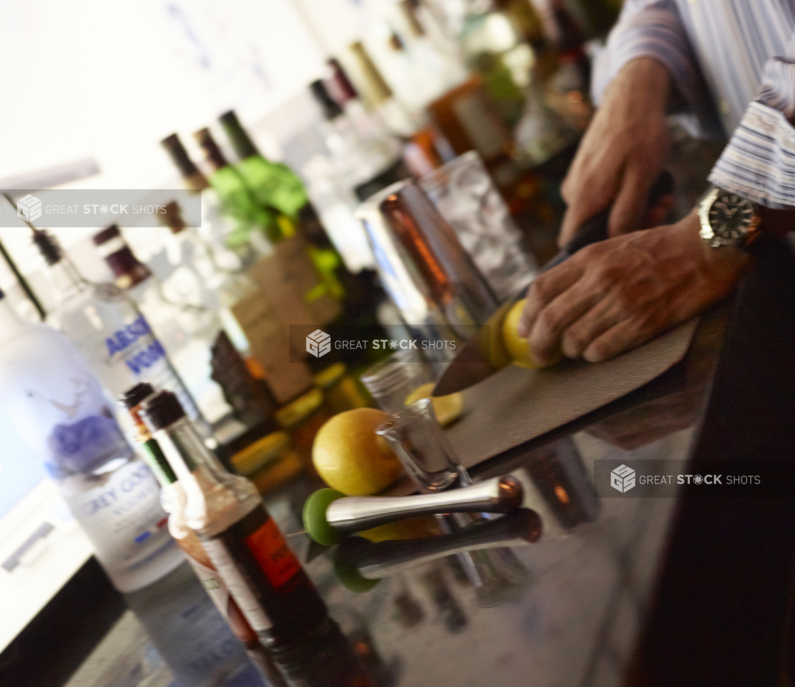 Bartender Cutting Lemons for Cocktail Garnish Surrounded by Several Bottles of Assorted Alcohol at a Bar Counter in a Restaurant Setting