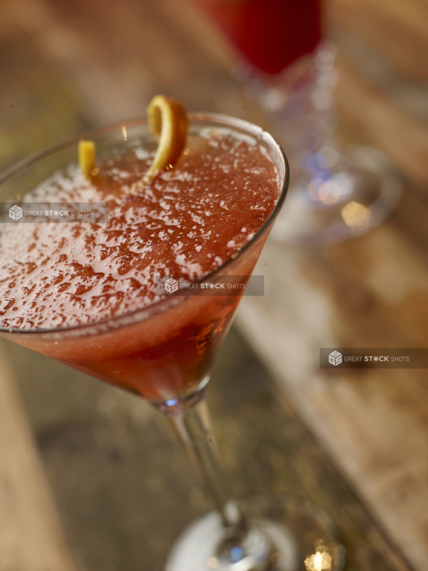 Close Up of a Martini Glass of Cosmopolitain with Crushed Ice and an Orange Peel Garnish on an Aged Wooden Surface