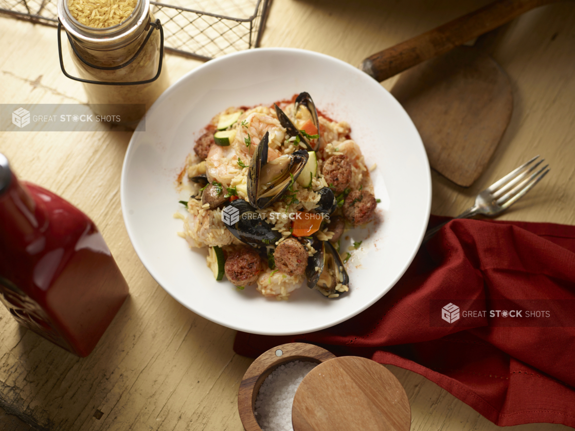 Overhead View of a Round White Dish of Seafood Paella on a White Painted Wooden Table with a Red Napkin and Wooden and Glass Kitchen Items