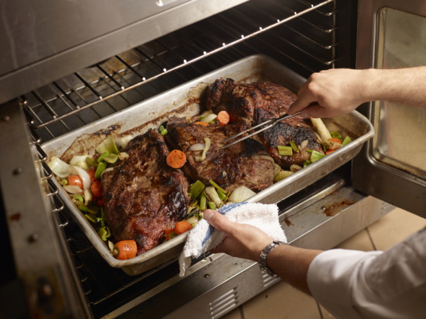 Chef Piercing a Large Roast Beef Chunk in a Roasting Pan with Assorted Vegetables in a Restaurant Kitchen Setting